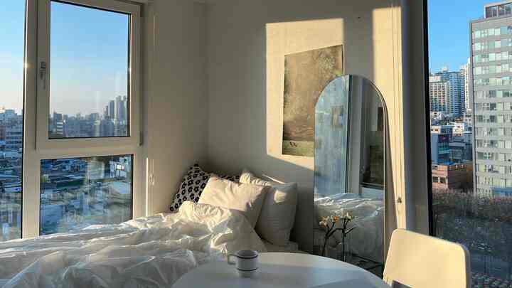 自然光が差し込む白を基調とした寝室、大きな窓とアーチ型全身鏡、白い寝具と丸いテーブルが配置されたミニマルな空間