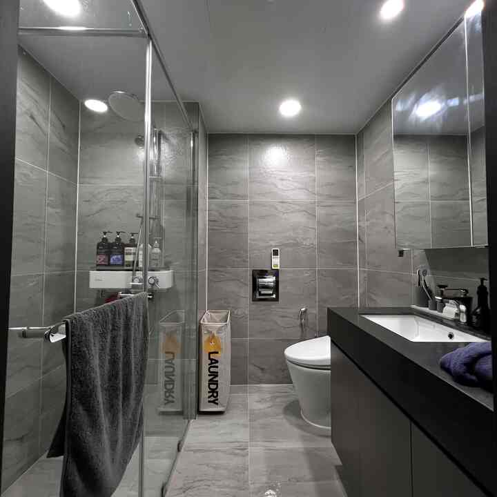 グレーのタイル壁と黒の洗面台があるモダンなバスルーム、狭い空間にランドリーバスケットが配置された清潔感のあるインテリア