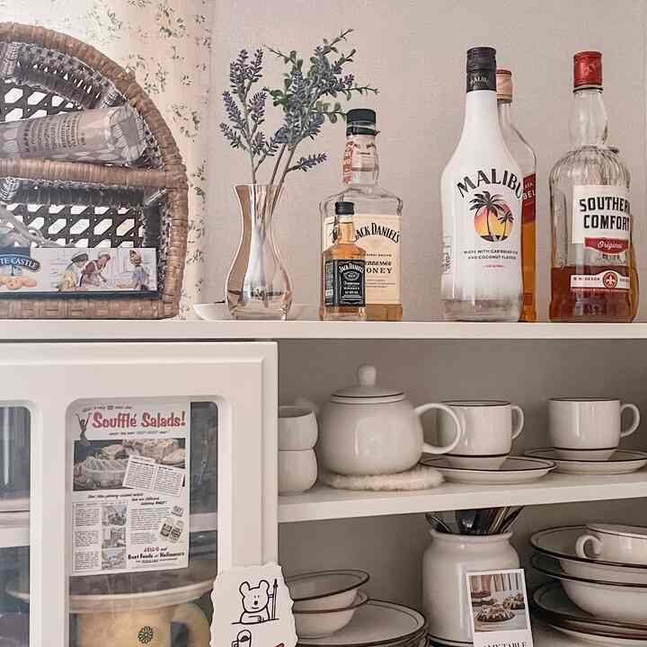白を基調としたキッチン収納の上にナチュラルなラベンダーの造花と複数の酒瓶が並ぶ、雰囲気のあるホームバー空間