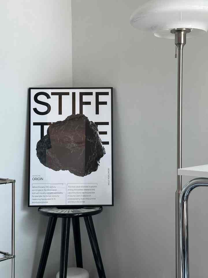 黒いスツールの上に置かれたポスターとシルバーのイケアトルビンフロアランプが配置されたシンプルでモダンなコーナースペース