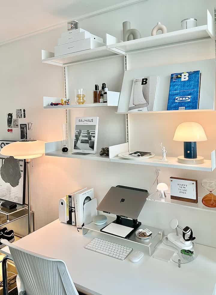 白とモノトーンのトーンのホームオフィス、デスクとレアロウの棚に整然と並べられた小物と照明が配置されたシンプルな雰囲気のインテリア