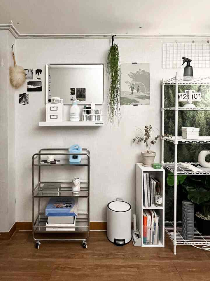 白い壁と木目調の床が調和する狭くコンパクトな部屋、移動式トロリーの化粧台と壁掛け棚、植物が配置された清潔感のあるインテリア