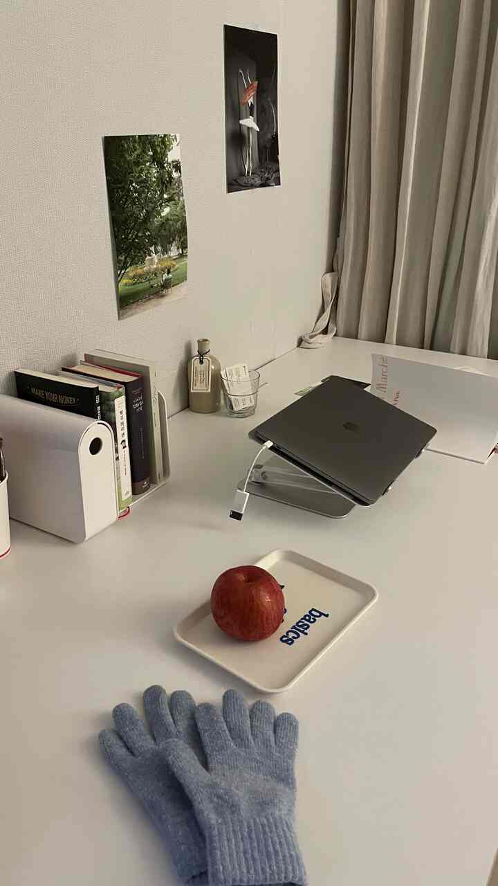 ホワイトの机とベージュのカーテンがあるホームオフィス空間、ノートパソコンスタンドと本棚の小物が配置されたシンプルな作業スペース