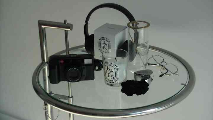 シルバーのクロームフレームを持つ丸いサイドテーブルに黒いカメラ、ヘッドホン、キャンドルが置かれたシンプルでモダンなリビング空間のインテリア