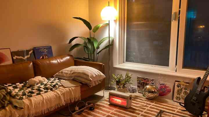 暖かいブラウン調のリビング、レザーソファとブランケット、電気ヒーター、フロアランプ、ギターが配置された居心地の良い空間