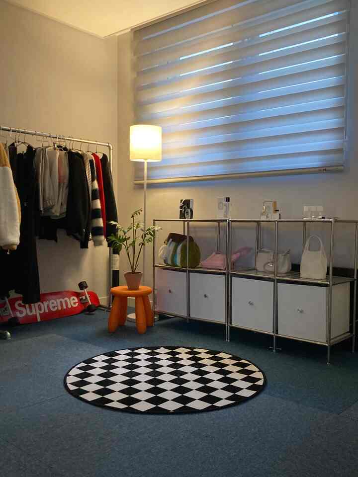 白い壁と青いカーペットのウォークインクローゼット、中央にチェッカーボードパターンの円形ラグとオレンジのスツール、整理された棚と洋服掛けが配置された印象的な空間