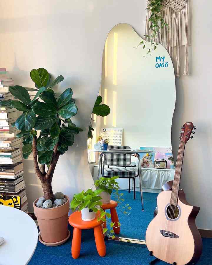 ベージュの壁と青いカーペットのワンルーム、変形全身鏡と植物、ギター、オレンジ色のスツールが配置された雰囲気のあるインテリア