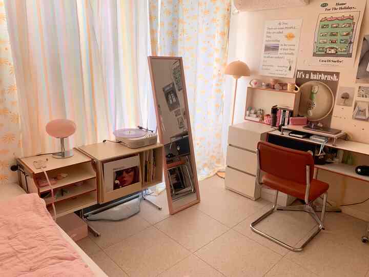 ベージュとアイボリーのトーンの小さな寝室、ピンクのダイニングチェアと全身鏡、LPターンテーブル、ポスターが並ぶ居心地の良い作業スペース