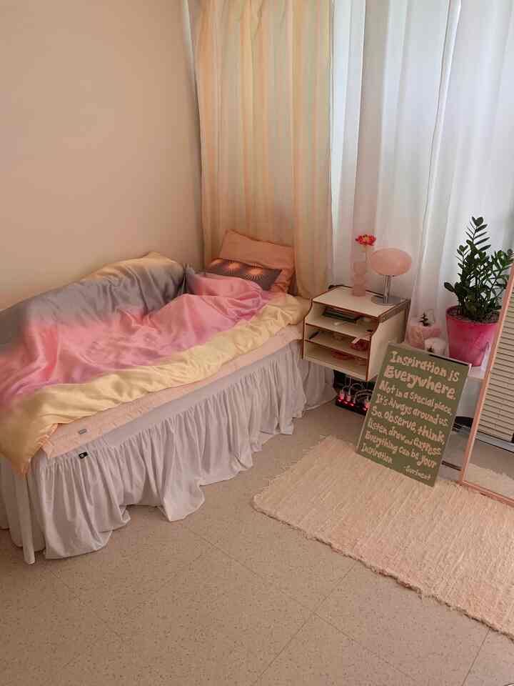 パステルトーンの寝室、淡いピンクの寝具と白いナイトテーブル、ラグ、植物が配置された雰囲気のあるインテリア