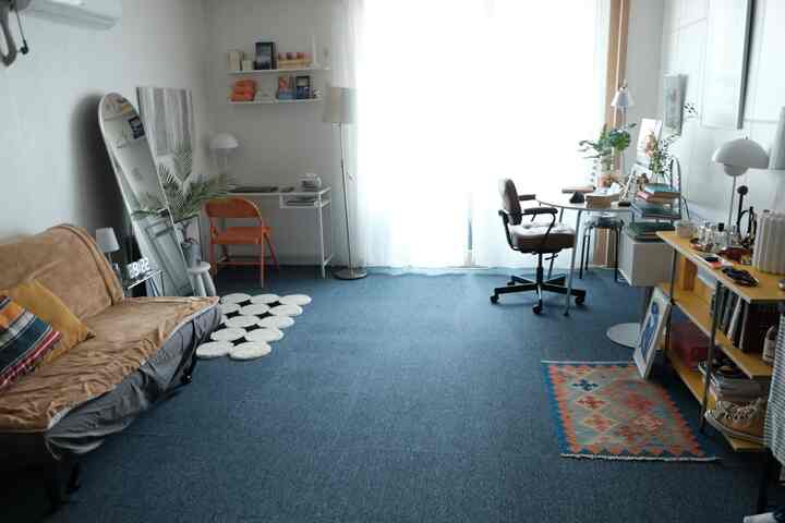 ブルーのカーペットが敷かれたワンルームの空間で、左側にブラウンのソファと折りたたみ椅子、右側にデスクとオフィスチェアが配置されたホームオフィススペース