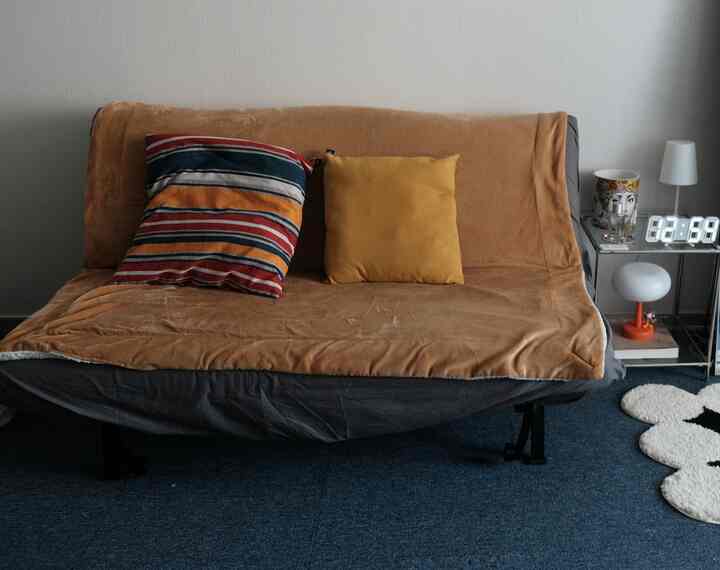 ブラウンとブルーのトーンのワンルーム空間、折りたたみ式ソファベッドとクッション、小さなナイトテーブルと時計が配置された雰囲気のあるインテリア