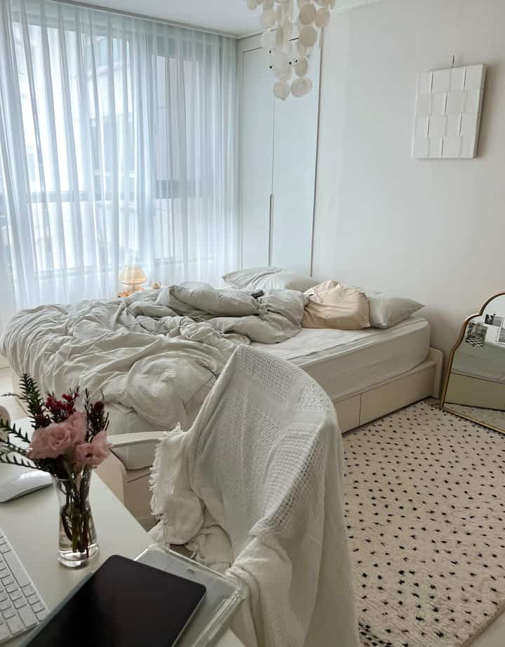 柔らかなアイボリーとベージュの色調のモダンなワンルーム寝室、窓際に白いカーテンとブランケット、整理されたデスクのある居心地の良い空間