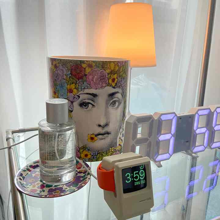 透明なガラス瓶と花柄の皿が置かれたシルバーのメタルナイトテーブルに、フォルナセッティのキャンドル陶器と充電式LEDスタンドライトが配置されたナチュラルカラーの寝室