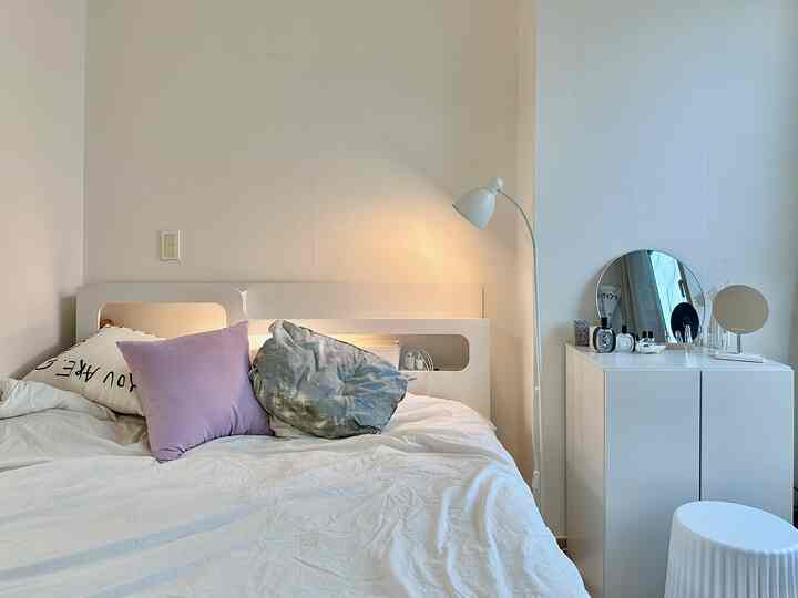 白とラベンダーの色調の寝室、クイーンサイズベッドと遮光カーテン、スツール、フロアランプが配置された雰囲気のあるモダンな空間