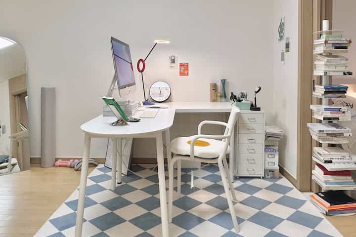 ホワイトとブルーのトーンのホームオフィス、L字型のデスクとチェック柄のラグ、本棚と整頓された収納が配置されたモダンな空間