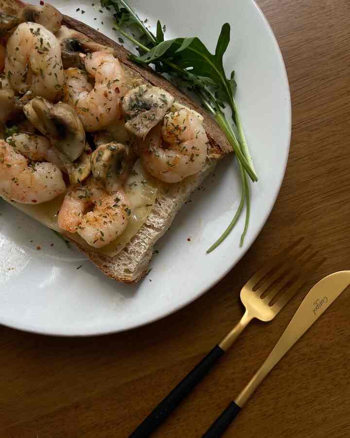木目調のテーブルの上に白い皿が置かれ、エビとキノコがのったパンのシンプルな食事が配置されている