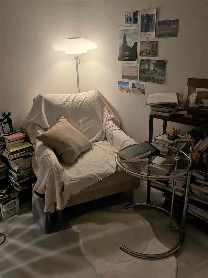 ナチュラルな色調の狭いコーナールーム、白いカバーのアームチェアとフロアランプ、丸いサイドテーブルが配置された居心地の良い読書スペース