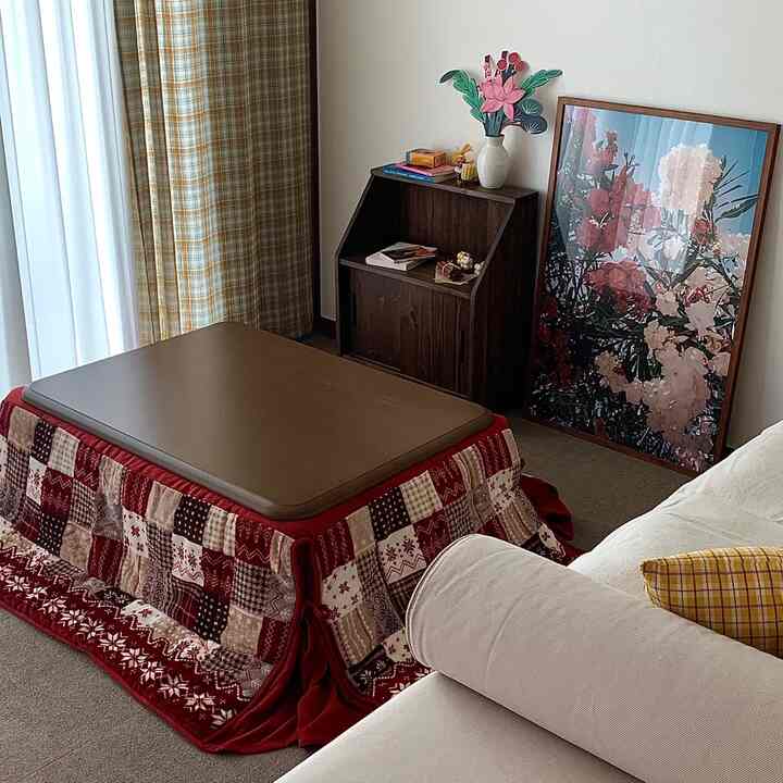 ベージュと木目調が調和するリビング、中央にこたつとチェック柄カーペット、右側にクリーム色のソファと花柄の額装アートが配置された暖かい冬のインテリア