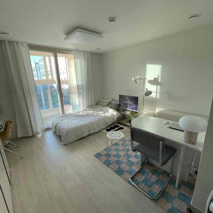 白とベージュのトーンのワンルーム、ベッドとデスク、ラグが配置されたシンプルモダンで落ち着いた雰囲気のインテリア