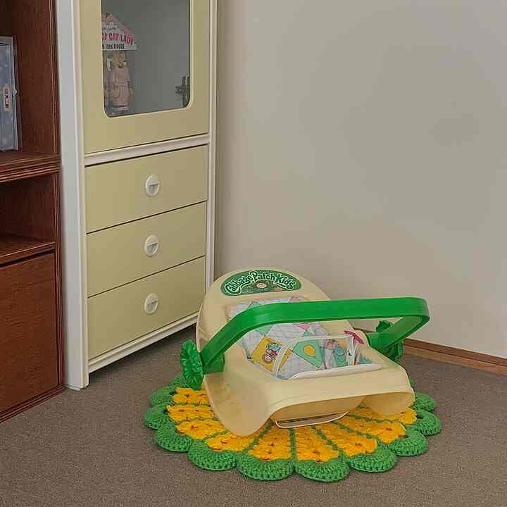 ベージュとグリーンの色調の小さな空間に、ヴィンテージ風のキャビネットとキャベツ人形のバウンサー、丸い編み物カーペットが配された温かみのある赤ちゃん部屋