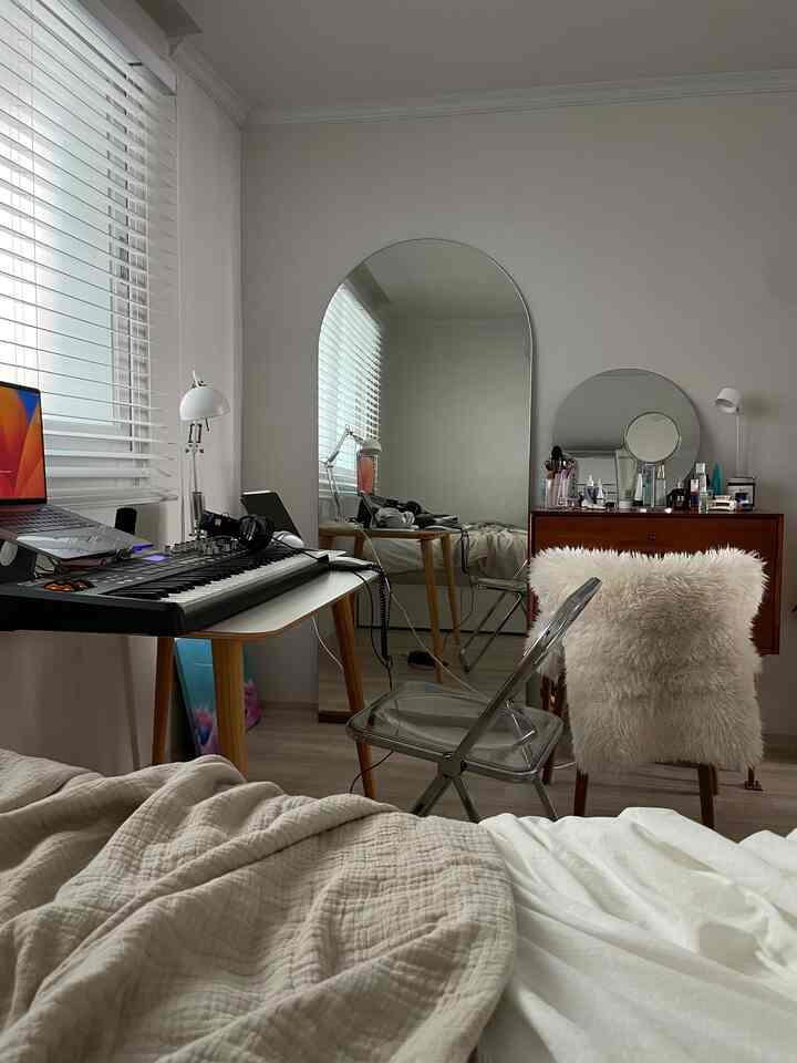 ベージュとホワイトのトーンの寝室、デスク、透明な椅子、2つの鏡と化粧台が配置された落ち着いた空間