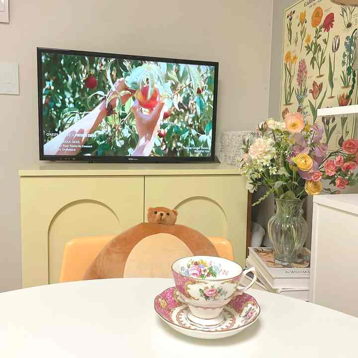 パステル調のダイニング、黄の装飾キャビネットと白いダイニングテーブルにビンテージティーカップが置かれ、花のポスターと花瓶が配された雰囲気のあるインテリア