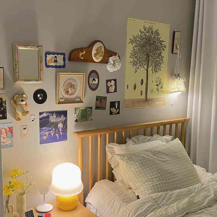 パステル調の寝室、木製ベッドとナイトテーブルのランプ、ヴィンテージの額やポスターが配置された落ち着いた空間