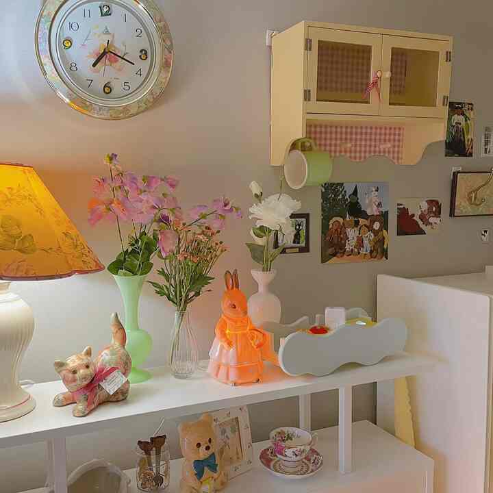 暖かいベージュ色の壁の寝室空間、ヴィンテージのフラワーパターンのテーブルランプとオレンジのウサギの照明、多様なヴィンテージの小物と額縁が配置された雰囲気のあるインテリア