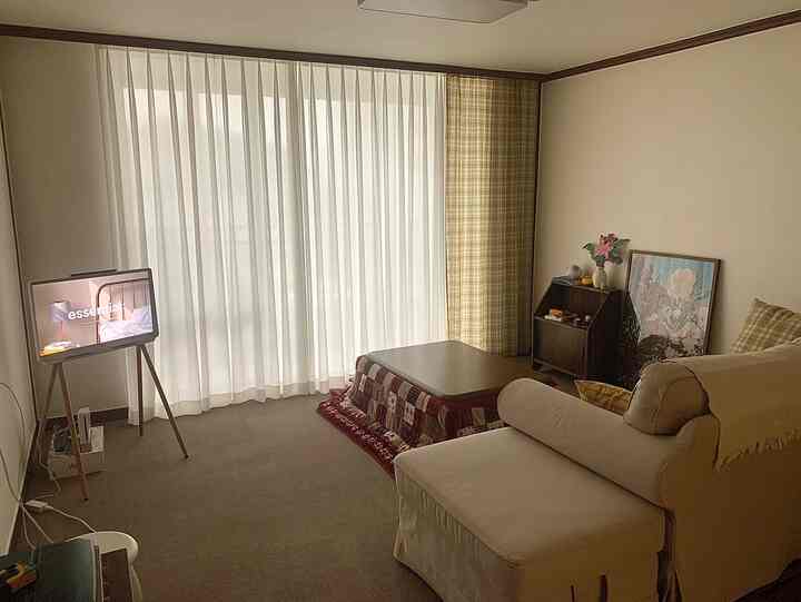 柔らかなベージュ色調のリビングルーム、大きな窓にカーテンが掛けられ、ソファとこたつテーブルが配置された雰囲気のあるインテリア