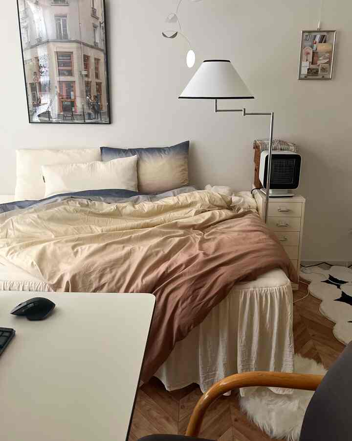 ホワイトとナチュラルカラーのワンルーム寝室、グラデーションの寝具とベッドスカート、ホームオフィスデスクが配置された雰囲気のあるインテリア