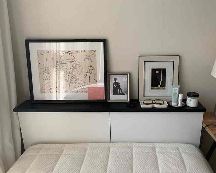 ホワイトの寝具とナチュラルトーンの壁紙がある寝室、収納棚の上の黒い棚に額縁や小物が置かれた落ち着いた空間