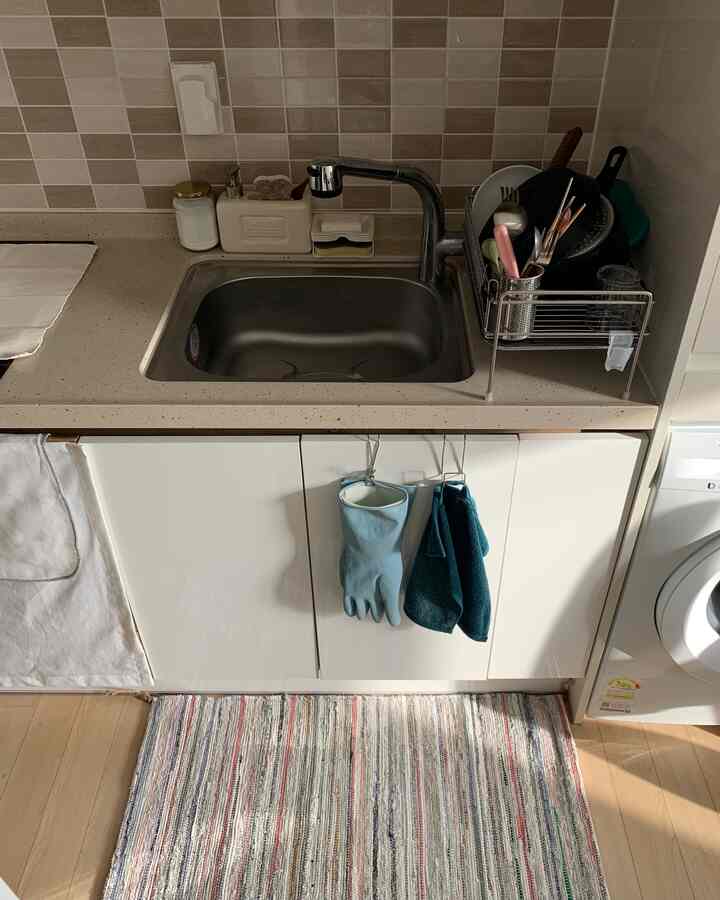 白い下部キャビネットとステンレス製のシンク、食器乾燥ラックが配置された清潔感のあるキッチン空間、床にはストライプのラグが敷かれている