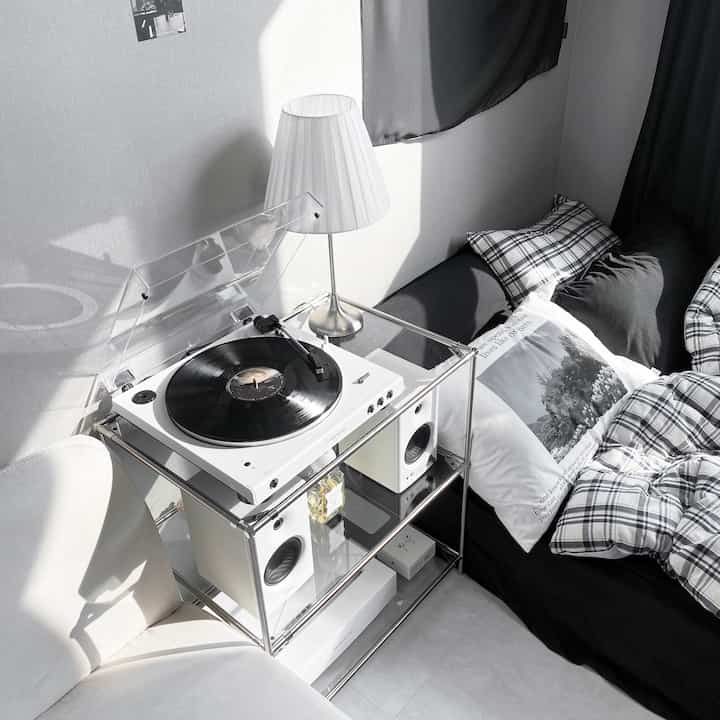 モノトーンの小さなリビング、黒いソファにチェックの枕があり、白いターンテーブルと棚、照明が配置された洗練されたモダンなインテリア