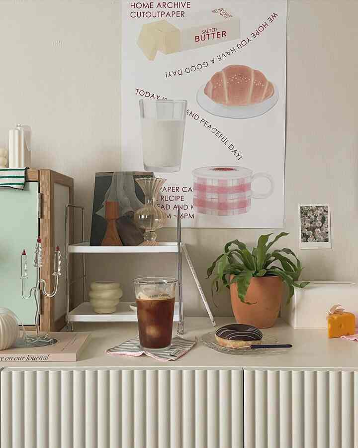 ナチュラルな色調のリビング空間、中央の観葉植物とグラスカップに入ったアイスコーヒー、ドーナツが置かれたホワイトキャビネット、壁のアートが調和した雰囲気のあるカフェ風インテリア