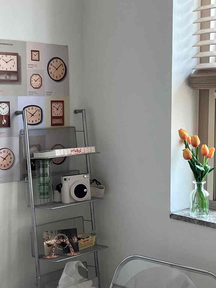 白とグレーのトーンの部屋、窓辺の棚にオレンジのチューリップ花瓶と鉄製のラダーシェルフが配置されたモダンで居心地の良い空間