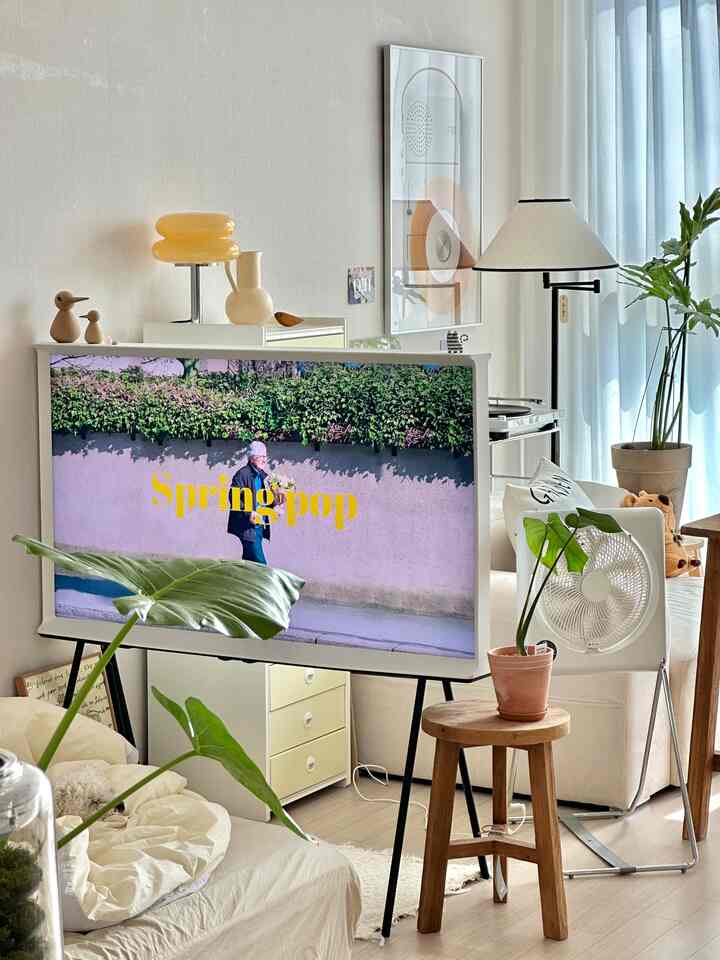 白と木目調を基調としたワンルームのリビング、大きなテレビとベージュのソファ、植物が配されたナチュラルな空間