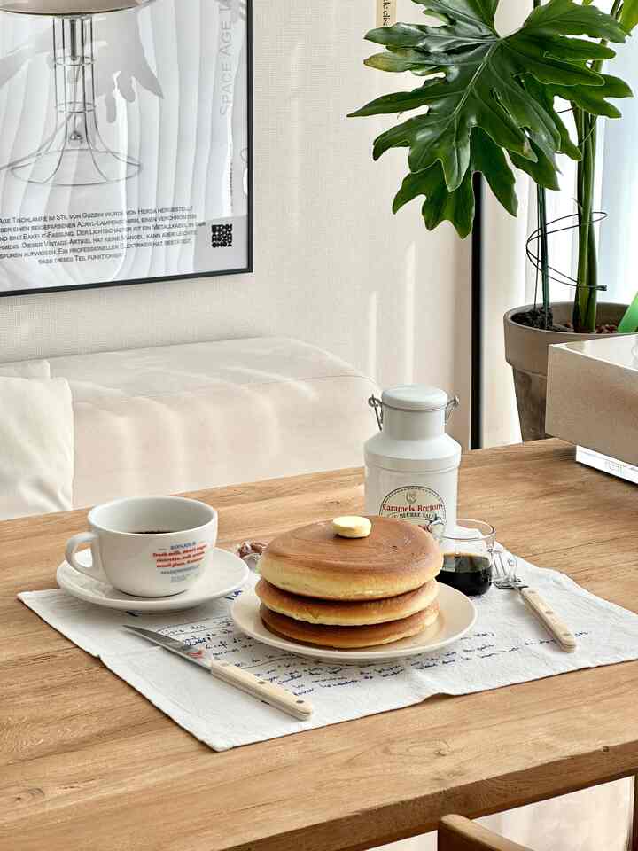 ナチュラルなブラウンとホワイトトーンのリビングダイニング空間、木製のダイニングテーブルにホームカフェ風のパンケーキとコーヒーセットが配置された落ち着いた雰囲気のインテリア