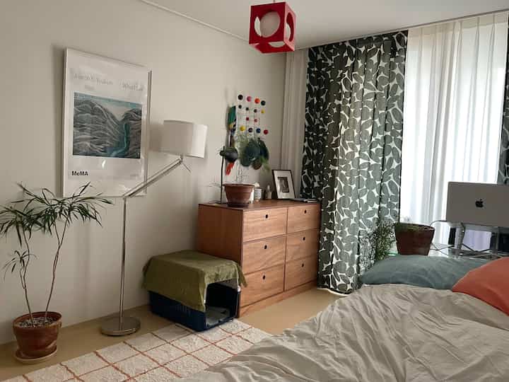 ホワイトとウッドトーンが調和する寝室、木製のドレッサーとフロアランプ、柄のカーテン、観葉植物が配置された居心地の良い空間