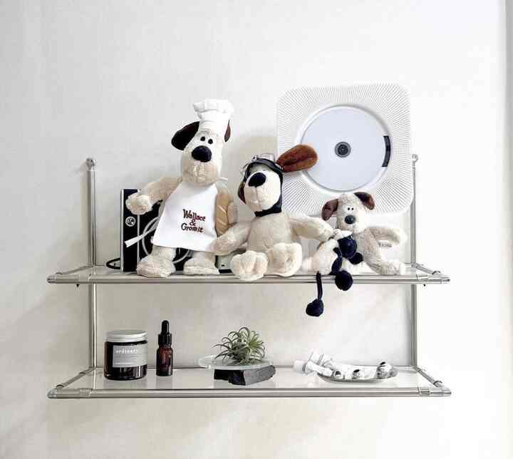 白を基調とした壁にステンレス製の壁掛け棚が設置され、上段にはウォレスとグルミットのぬいぐるみとCDプレーヤー、下段には植物とオブジェが置かれたすっきりとしたインテリア