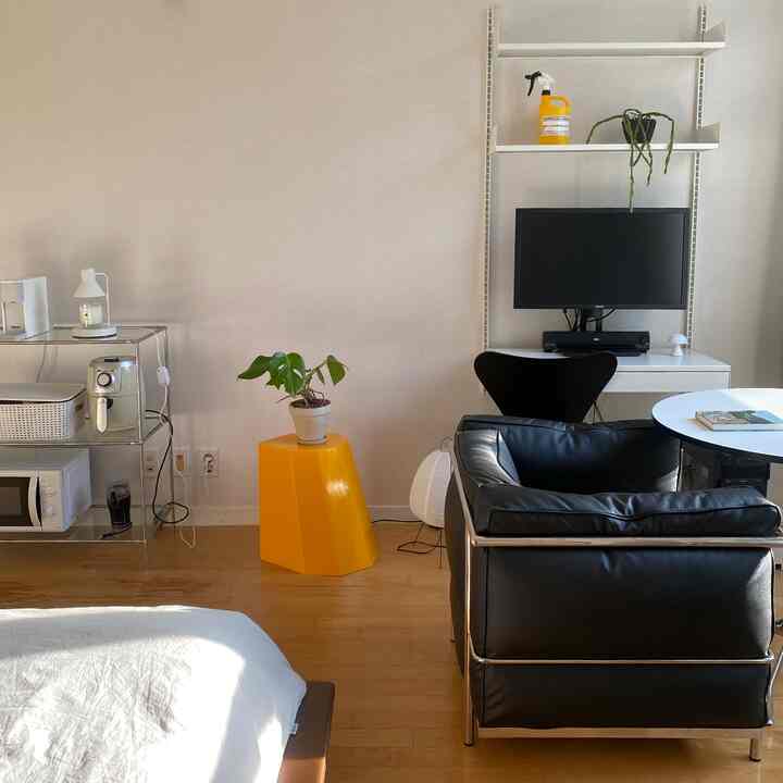 白と茶色のトーンの小さなワンルーム、黒いレザーのアームチェアと黄色のスツール、モニターデスクが配置されたモダンで居心地の良い空間