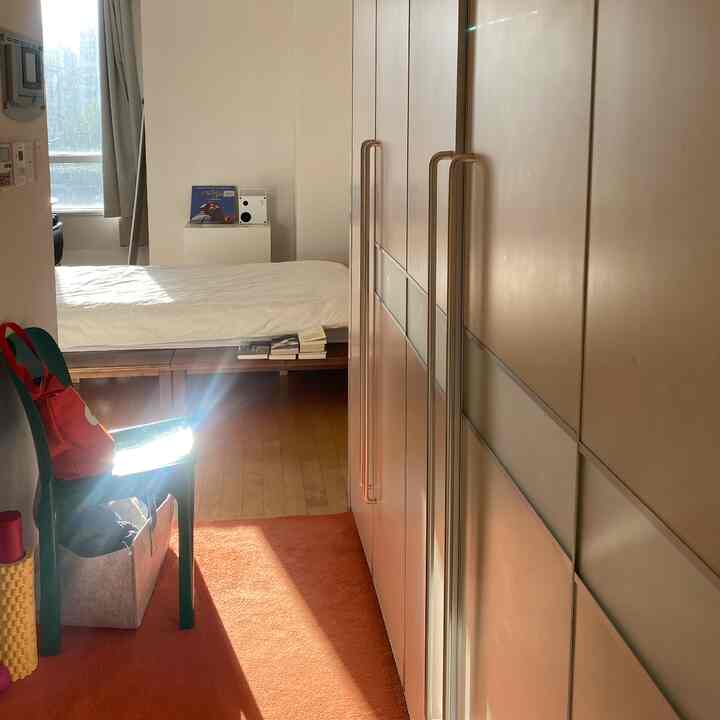 ベージュ調のワンルーム寝室、大きな収納棚が右側に配置され、オレンジのラグとベッドが調和した雰囲気のあるインテリア