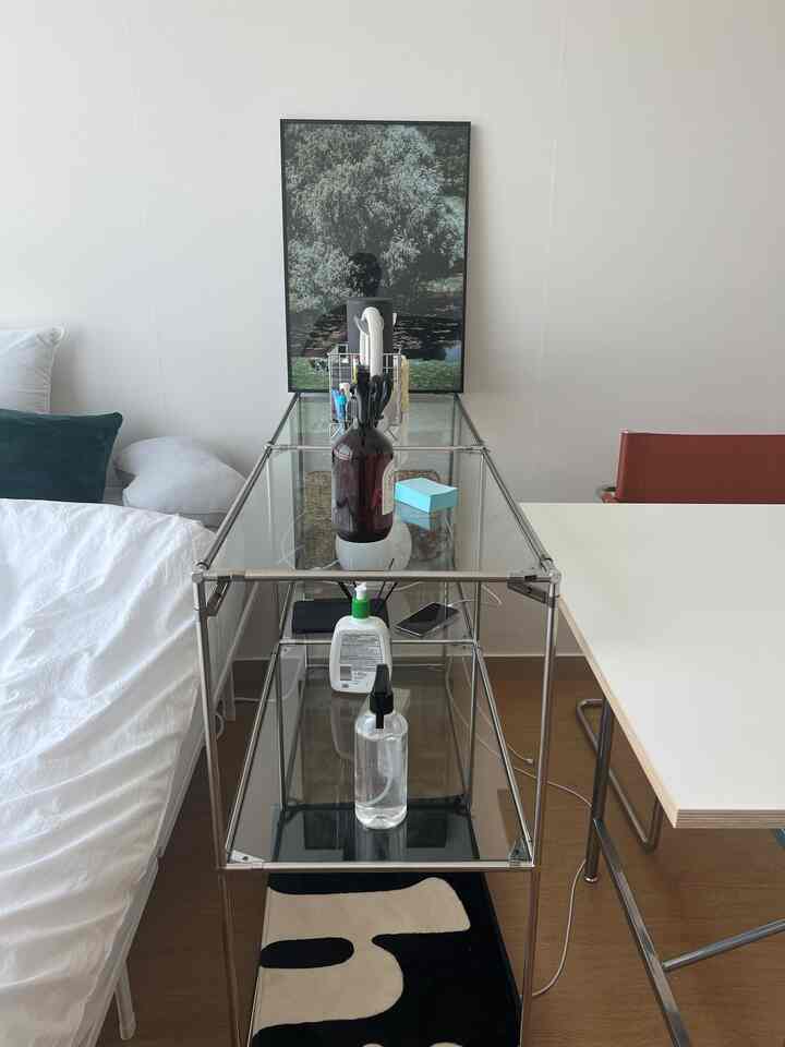 白い壁と木目調の床のワンルーム、透明ガラスの棚、ベッド、白い机と茶色の椅子が配置されたモダンで清潔感のある空間