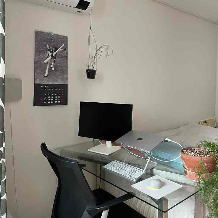 透明なガラステーブル、モニター、ノートパソコン台が配置された白を基調とした寝室、清潔で開放感のあるホームオフィス環境