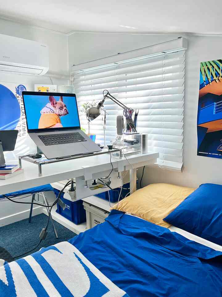 白い壁とブラインドのあるロフト、青い寝具とデスク上のノートパソコンとランプが配置された雰囲気のあるホームオフィス