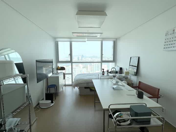 白と木目調を基調としたコンパクトなワンルーム、シンプルで整ったホームオフィススペースに机とベッドが配置されている