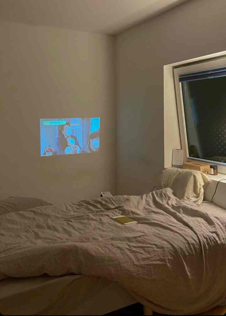 白い壁と低い天井のある狭い寝室、寝具とコンパクトなミニプロジェクターが配置された落ち着いた空間