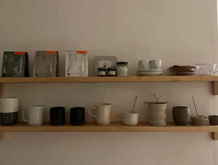 白い壁とベージュの木製棚がある玄関、陶器のカップやコーヒー豆、お香の小物が整然と配置された温かみのある空間