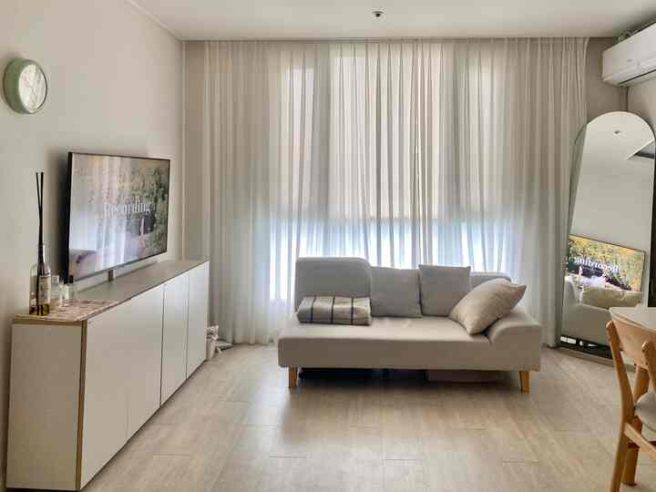 白を基調とした縦長のリビング、中央にソファ、壁にテレビ台とカーテンが配置されたシンプルな空間