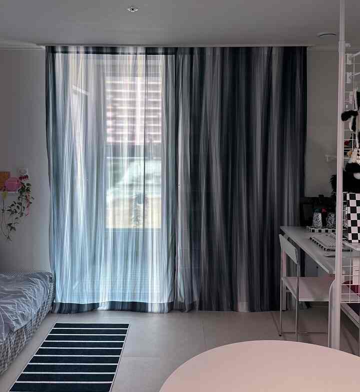 白とグレーのトーンのワンルーム寝室、窓に設置されたブラインドとカーテンが落ち着いた雰囲気を作るシンプルな空間