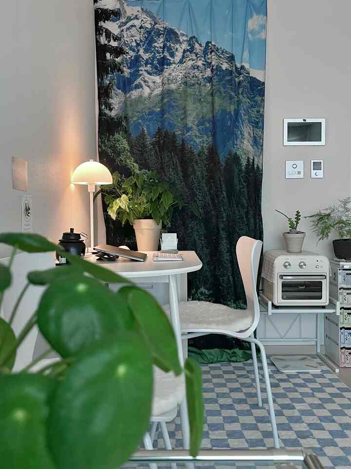 ナチュラルなトーンのホームオフィス空間、白いデスクと椅子、テーブルランプ、緑の植物、自然の風景が描かれたカーテンが配置された落ち着いたワークスペース